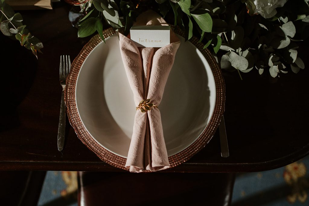 A rolled and folded dusky pink textured napkin is held together by a gold napkin ring decorated with leaves. The napkin sits on top of a cream charger plate with a brown textured rim. Above the plate is a small, minimalistic name card with calligraphy writing and green foliage decorates the centre of the table.