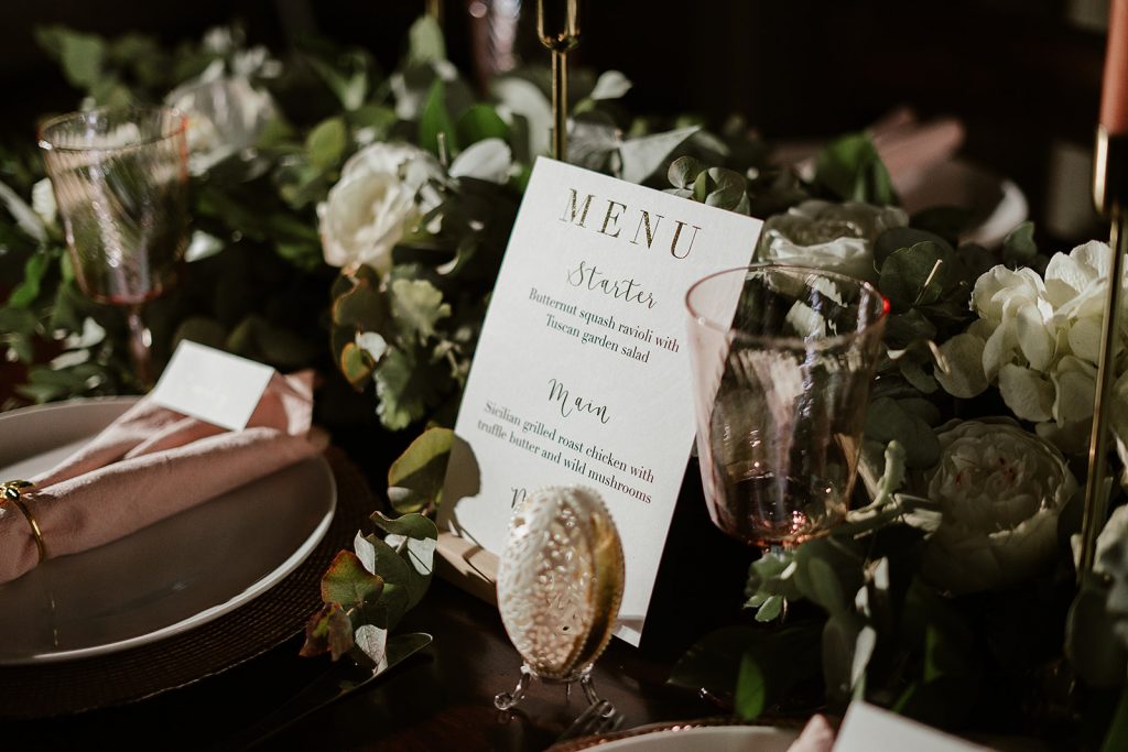 A small contemporary menu card with plain fonts and calligraphy writing is nestled amongst a mass of greenery on an intimate wedding tablescape. Pops of white flowers intersperse the greenery and a delicate, hand carved egg sits in front of the menu as a gorgeous wedding centrepiece.