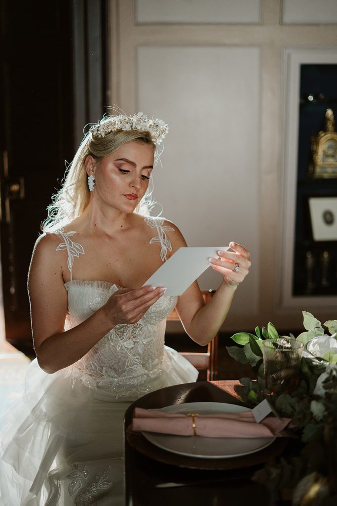 A bride sits at a wedding table at The HAC in London. She wears an embroidered, shimmery couture wedding dress with thin straps which have embroidered lead motifs attached. She wears an extravagent silver and pearl headpiece and dangly white flower earrings. She holds a wedding menu in her hands as she looks down reading it.