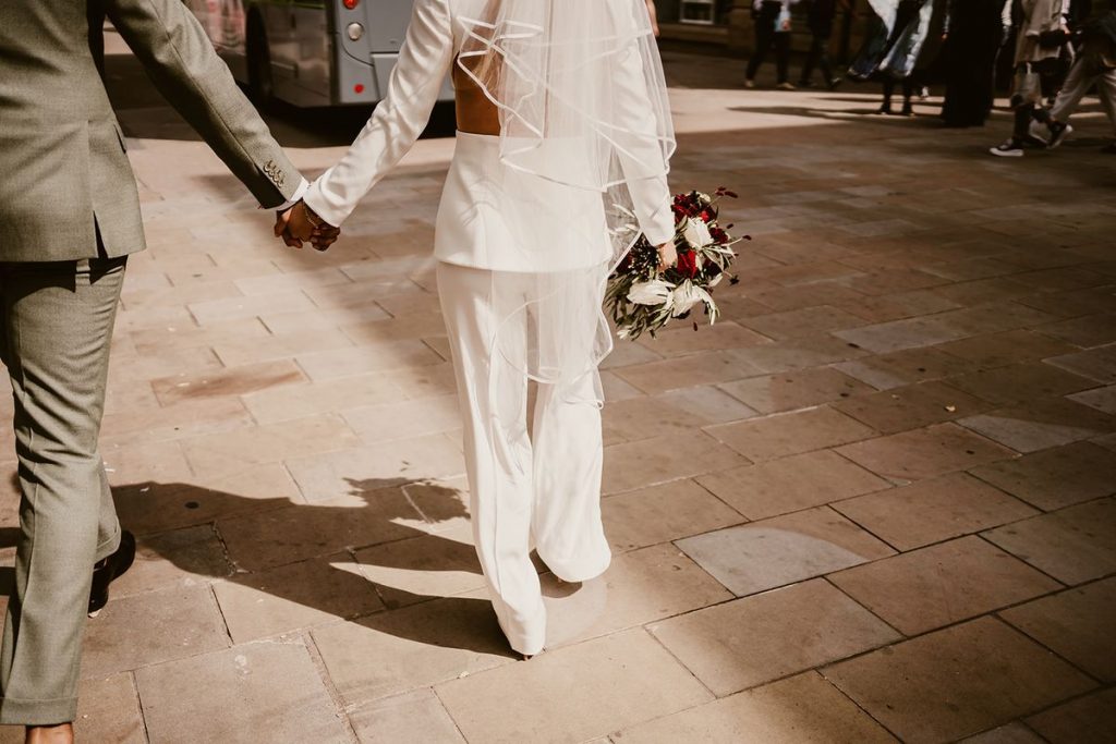 A very modern, chic bride wears a four piece wedding suit and walks with her new husband through a city street. You can see the back of the bride from the neck down and the side of the groom. The hold hands as they walk. In the bride's right hand is a small wedding bouquet of red and white flowers and green foliage.
