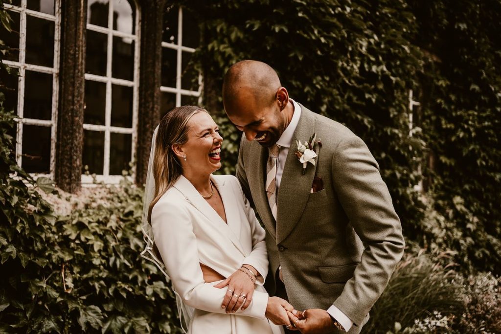 A bride and groom laugh hysterically with each other as they hold onto one another. The groom looks down towards the floor as he laughs, holding onto his bride's hand tightly. The bride is extremely stylish, wearing a contemporary white wedding suit and a mid length veil She wears several pairs of small earrings, a simple chain necklace and lots of rings on all of her fingers.