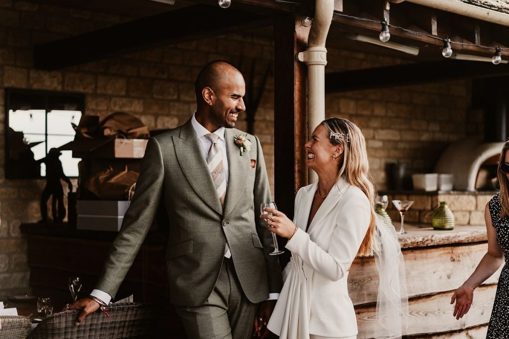 A newly married couple look at one another smiling broad smiles. The groom wears a smart suit with a tie and small button hole. The Bride wears a cream wedding suit. She looks chic and very stylish as she holds a glass of Champagne in one hand. She has a sparkling diamanté hair clip that reads 'Mrs B'.