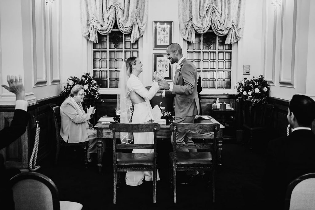 A bride and groom face one another and hold hands in a ceremony room at Oxford Registry Office moments before saying their vows to one another. The both have the look of utter adoration and genuine delight on their faces.