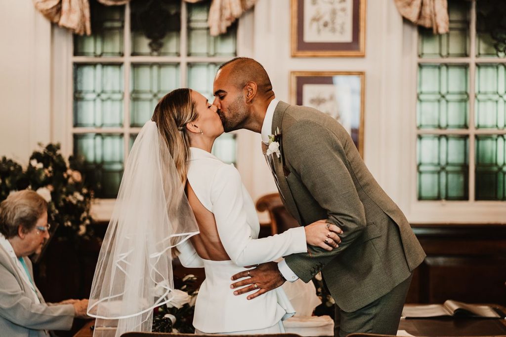 A bride and groom share their first kiss as newly weds during their civil ceremony in a registry office. The groom bounds down and forward to kiss his new wife, as she leans back slightly. The groom holds his bride gently around the waist and she holds onto his forearm. The bride wears a chic bridal suit with a cut out panelling section, exposing her back. She wears a mid length veil.