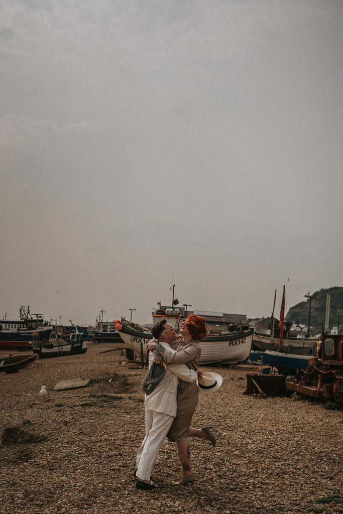 A queer couple are enjoying a beach elopement inspiration photo shoot. One of the lovers wears a white suit with a black vest underneath and a white hat with a black band. The other lover wears a vintage style Old Hollywood glamour under the knee length dress with a long pearl necklace. The couple are very much in love showing adoration for one another.