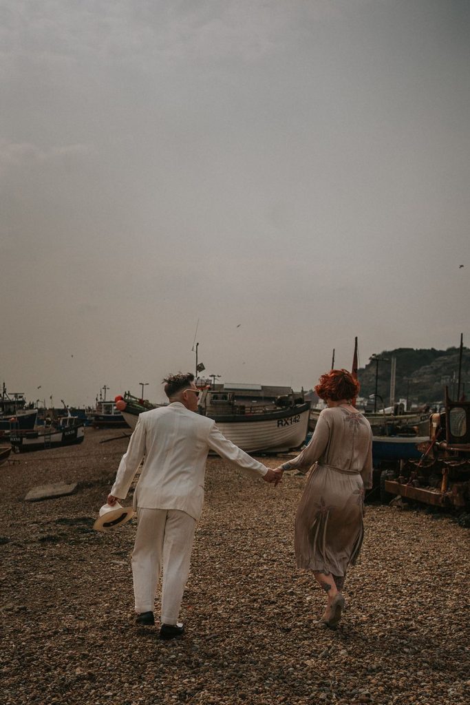 A queer couple are enjoying a beach elopement inspiration photo shoot. One of the lovers wears a white suit with a black vest underneath and a white hat with a black band. The other lover wears a vintage style Old Hollywood glamour under the knee length dress with a long pearl necklace. The couple are very much in love showing adoration for one another.