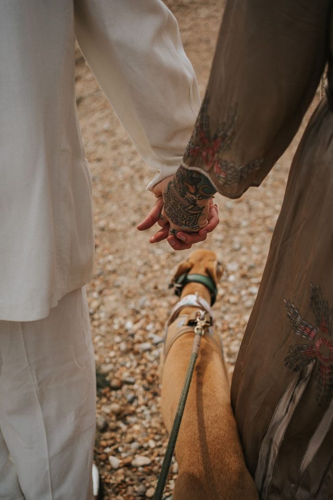 A queer couple are enjoying a beach elopement inspiration photo shoot. One of the lovers wears a white suit with a black vest underneath and a white hat with a black band. The other lover wears a vintage style Old Hollywood glamour under the knee length dress with a long pearl necklace. The couple are very much in love showing adoration for one another.
