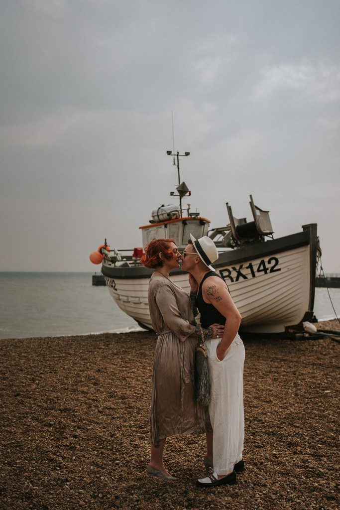 A queer couple are enjoying a beach elopement inspiration photo shoot. One of the lovers wears a white suit with a black vest underneath and a white hat with a black band. The other lover wears a vintage style Old Hollywood glamour under the knee length dress with a long pearl necklace. The couple are very much in love showing adoration for one another.