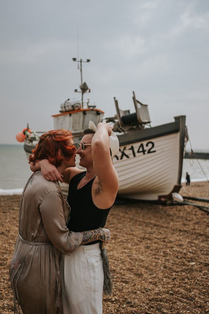 A queer couple are enjoying a beach elopement inspiration photo shoot. One of the lovers wears a white suit with a black vest underneath and a white hat with a black band. The other lover wears a vintage style Old Hollywood glamour under the knee length dress with a long pearl necklace. The couple are very much in love showing adoration for one another.