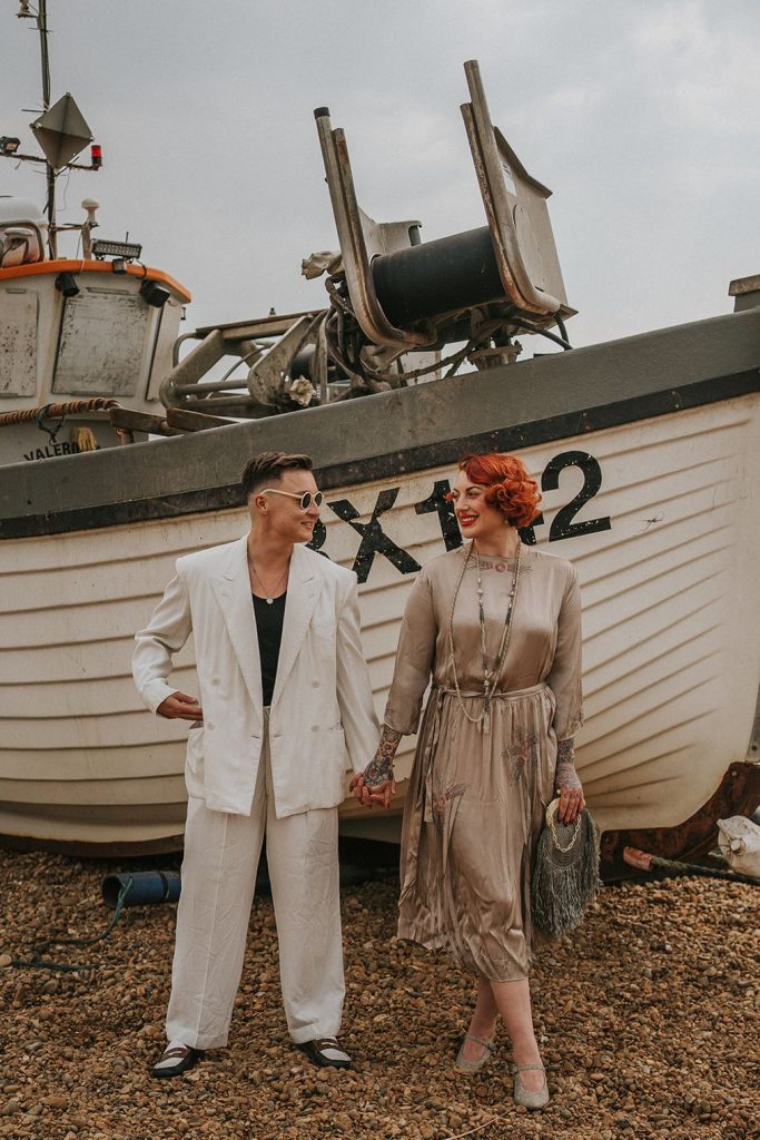 A queer couple are enjoying a beach elopement inspiration photo shoot. One of the lovers wears a white suit with a black vest underneath and a white hat with a black band. The other lover wears a vintage style Old Hollywood glamour under the knee length dress with a long pearl necklace. The couple are very much in love showing adoration for one another.