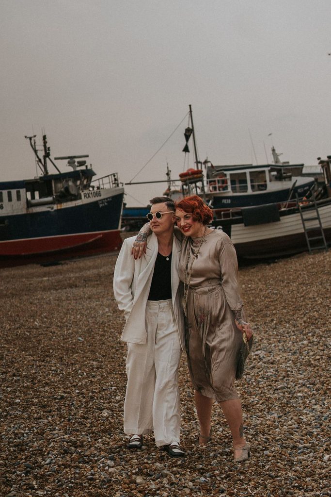 A queer couple are enjoying a beach elopement inspiration photo shoot. One of the lovers wears a white suit with a black vest underneath and a white hat with a black band. The other lover wears a vintage style Old Hollywood glamour under the knee length dress with a long pearl necklace. The couple are very much in love showing adoration for one another.