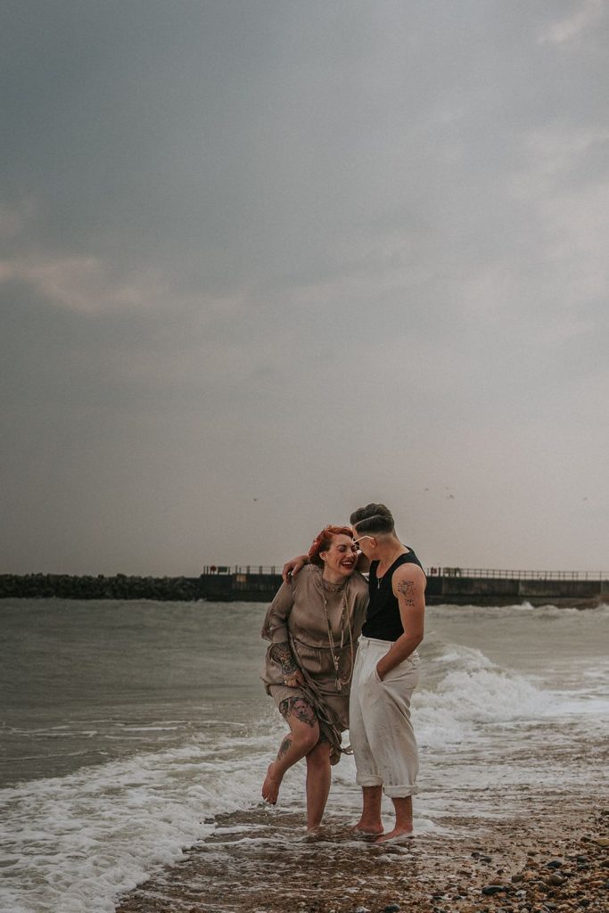 A queer couple are enjoying a beach elopement inspiration photo shoot. One of the lovers wears a white suit with a black vest underneath and a white hat with a black band. The other lover wears a vintage style Old Hollywood glamour under the knee length dress with a long pearl necklace. The couple are very much in love showing adoration for one another.