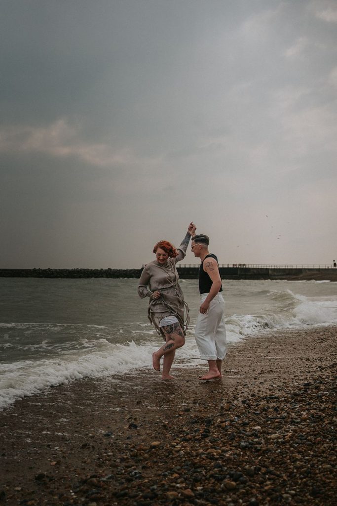 A queer couple are enjoying a beach elopement inspiration photo shoot. One of the lovers wears a white suit with a black vest underneath and a white hat with a black band. The other lover wears a vintage style Old Hollywood glamour under the knee length dress with a long pearl necklace. The couple are very much in love showing adoration for one another.