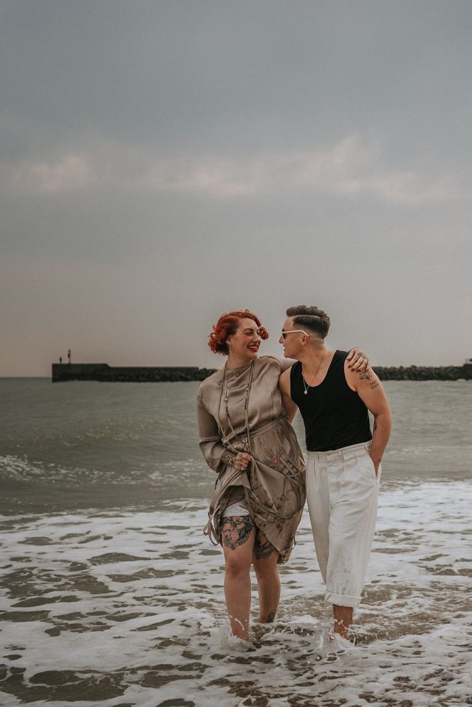 A queer couple are enjoying a beach elopement inspiration photo shoot. One of the lovers wears a white suit with a black vest underneath and a white hat with a black band. The other lover wears a vintage style Old Hollywood glamour under the knee length dress with a long pearl necklace. The couple are very much in love showing adoration for one another.