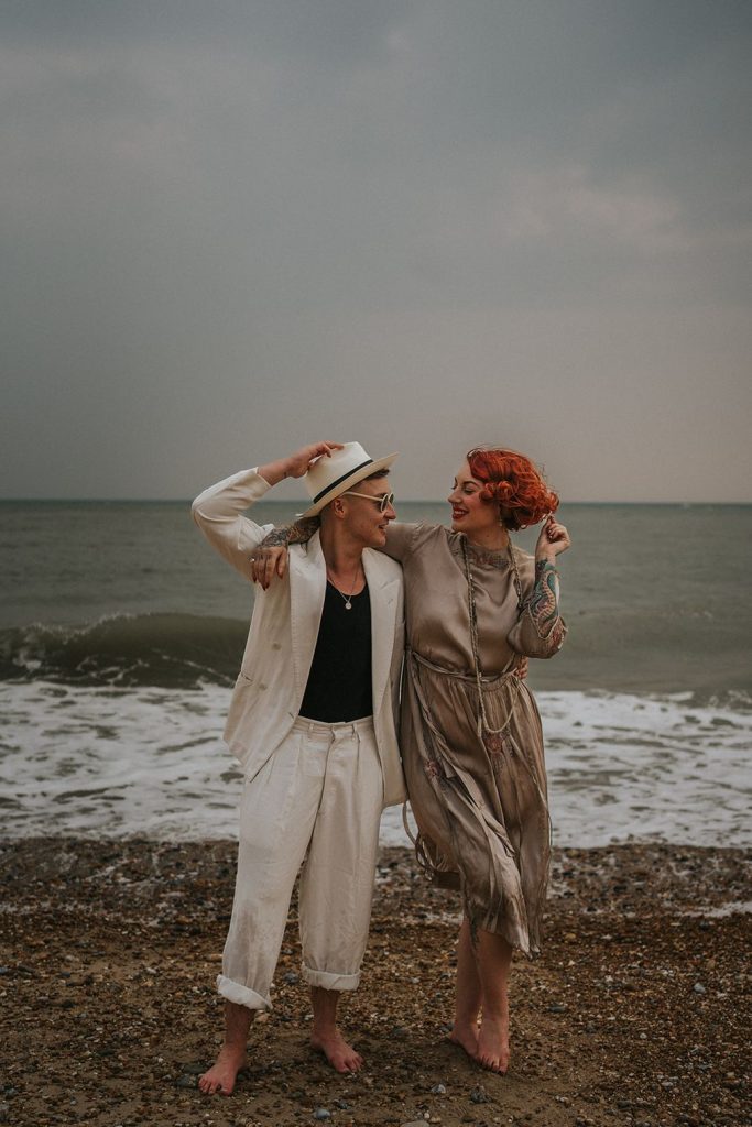 A queer couple are enjoying a beach elopement inspiration photo shoot. One of the lovers wears a white suit with a black vest underneath and a white hat with a black band. The other lover wears a vintage style Old Hollywood glamour under the knee length dress with a long pearl necklace. The couple are very much in love showing adoration for one another.