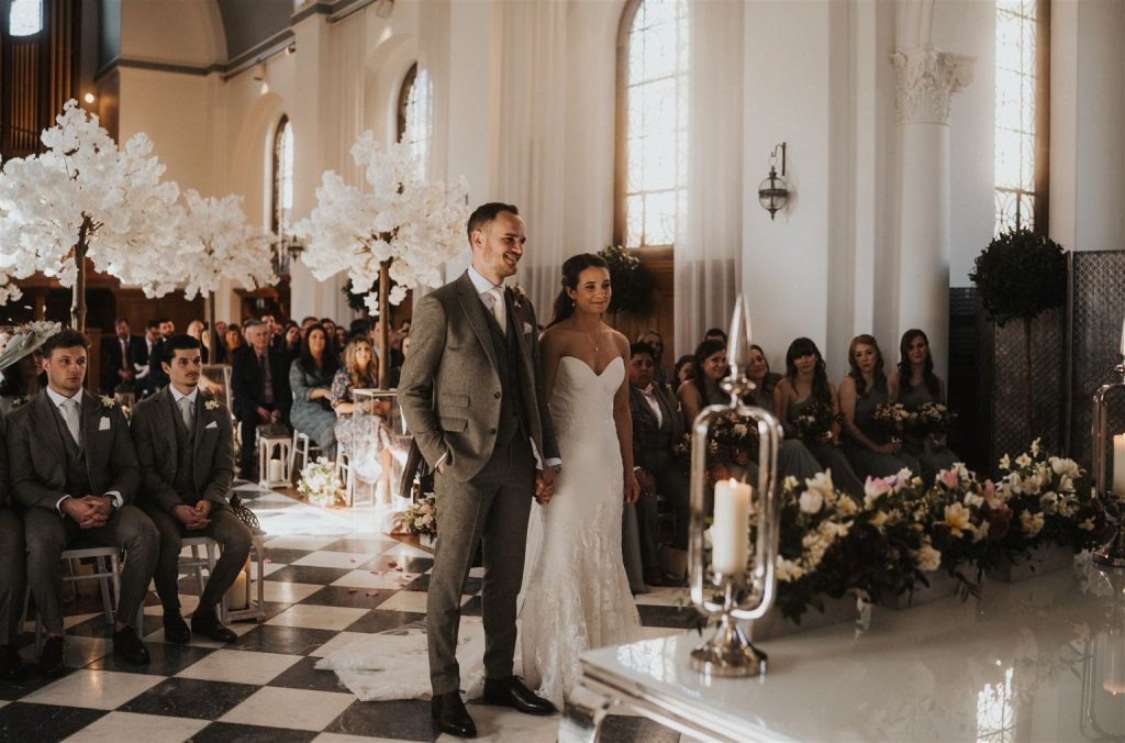 A bride and groom stand side by side holding hands, at the front of their civil ceremony room at Hawkstone Hall during their service. They both look forwards smiling. Behind them are rows and rows of guests, many of them smiling, and some looking quote emotional.