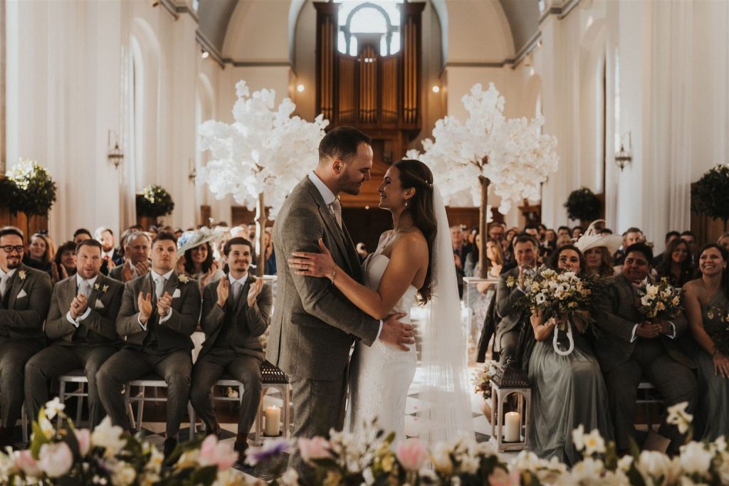 An almost newly wed couple stand in front of their guests in their wedding ceremony room facing one another and holding onto one another as they gaze longingly into one another's eyes. You can almost feel the love radiating from them as they look so intently at each other.