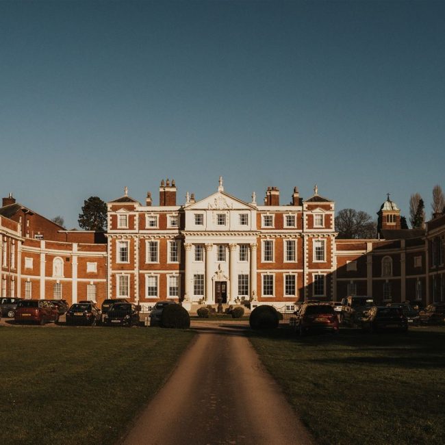 The back view of Hawkstone Hall wedding venue. It is a huge and very grand looking 18th century manor house with large open grass areas on which wedding guests are enjoying drinks and canapes underneath a deep blue spring sky.