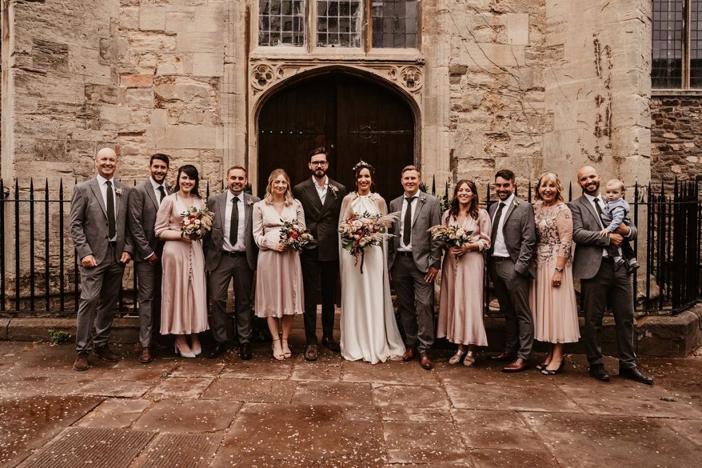 A wedding party stand lined up facing the camera for a group shot. They are standing alternative with men between women, The men are all smart wearign suits whilst the 4 bridesmiads wear dusky pink bridesmaids dresses. The groom stand proud and tall looking smart but casual in a suit but no tie with an open collar. The bride wears a halter neck Halfpenny London gown with a dramatic cape. She looks relaxed, natural and ethereal. All guests smile broadly as they look towards the camera.