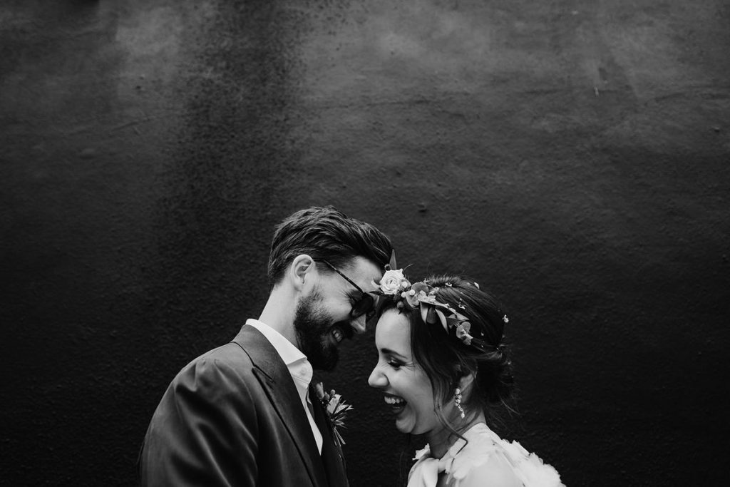A black and white image of newlyweds laughing with one another in an intimate moment. The faces and torsos are visible within the image. Both the bride and groom have large smiles as if laughing out loud. The groom has dark hair and glasses whilst the bride has dark hair worn on a loose up-do with fresh flower hair accessories.