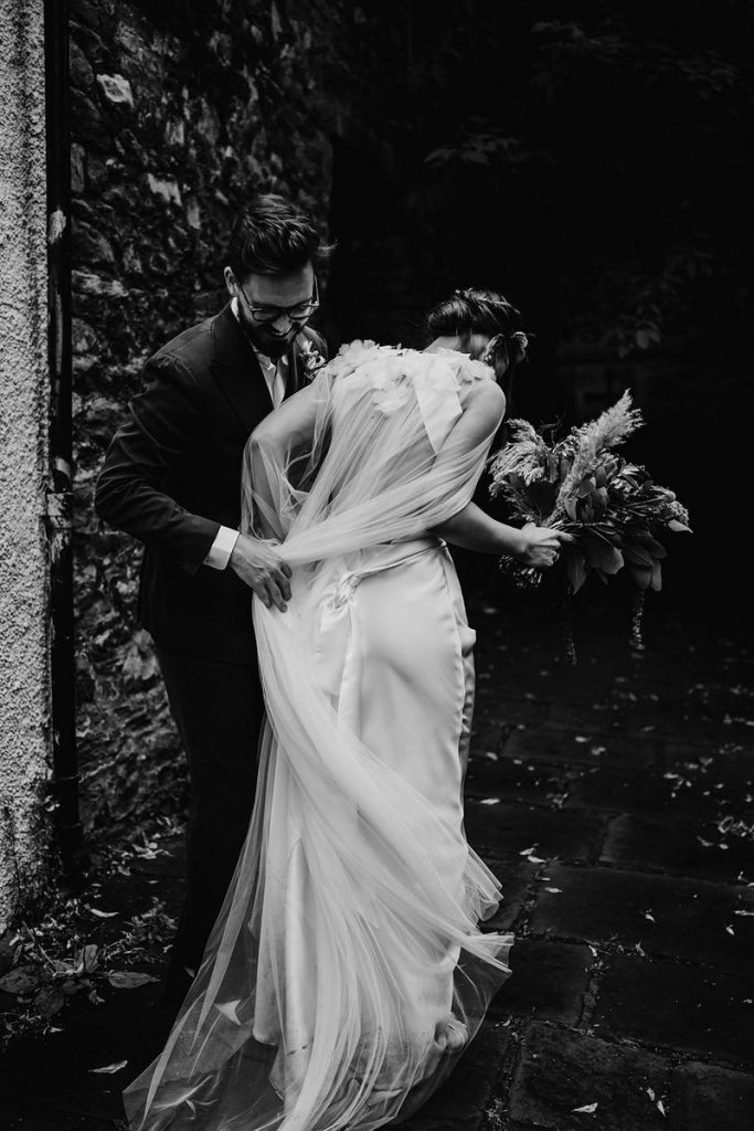 A black and white image of a bride with her new husband. The back of the bride is visible; she's moving her Halfpenny London wedding cape out from under her feet, as her husband helps. She wears an open backed Halfpenny London wedding dress.