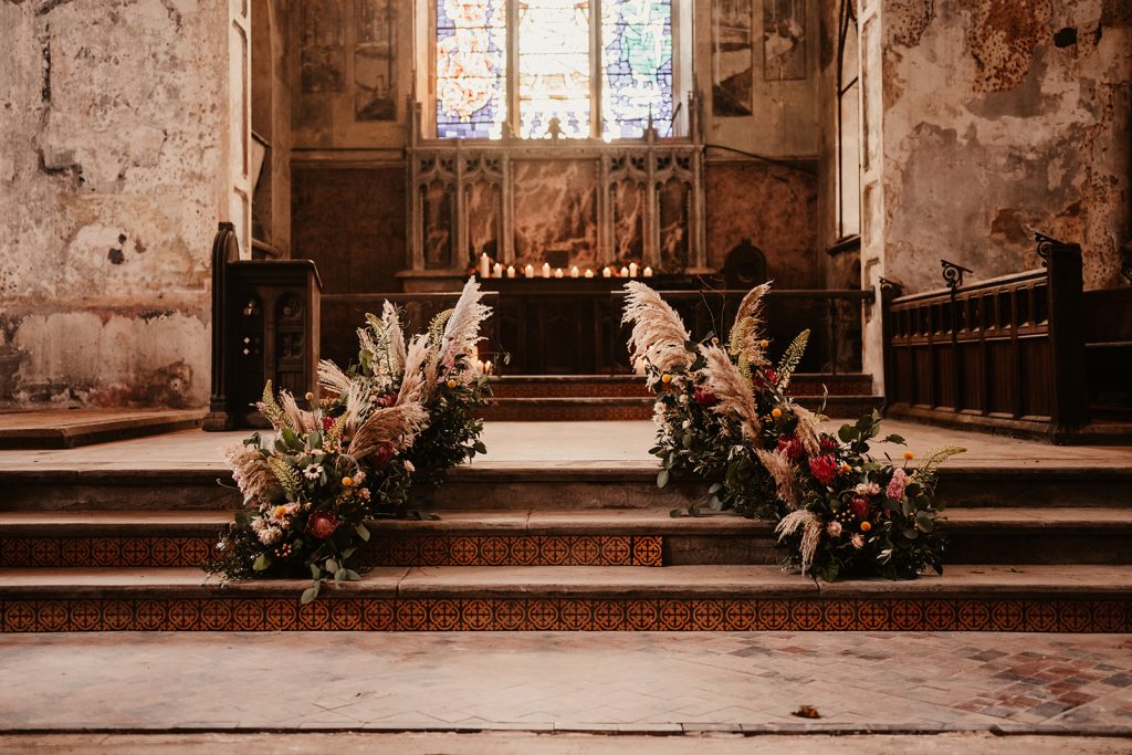 Exposed brick steps lead up to the alter / stage area of an old Church that is now used for wedding celebrations. The stairs are lined with large individual pampas and flower displays, with hints of pinks, reds and greens. In the background is a large and very impressive stained glass window.