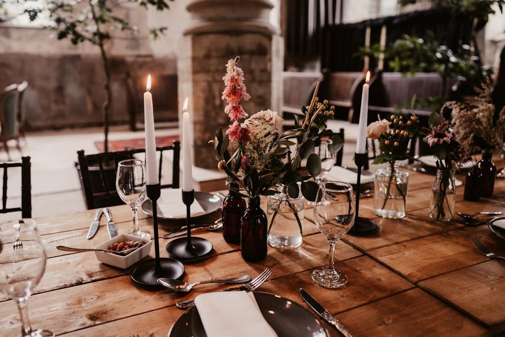 Long wooden tables are set out at The Mount Without in Bristol, ready for a wedding breakfast. The table is sparsely decorated, giving a simplistic and minimalist feel. Accents and hints of black sit alongside white candles, crcokery and glassware and hints of green and pink florals.