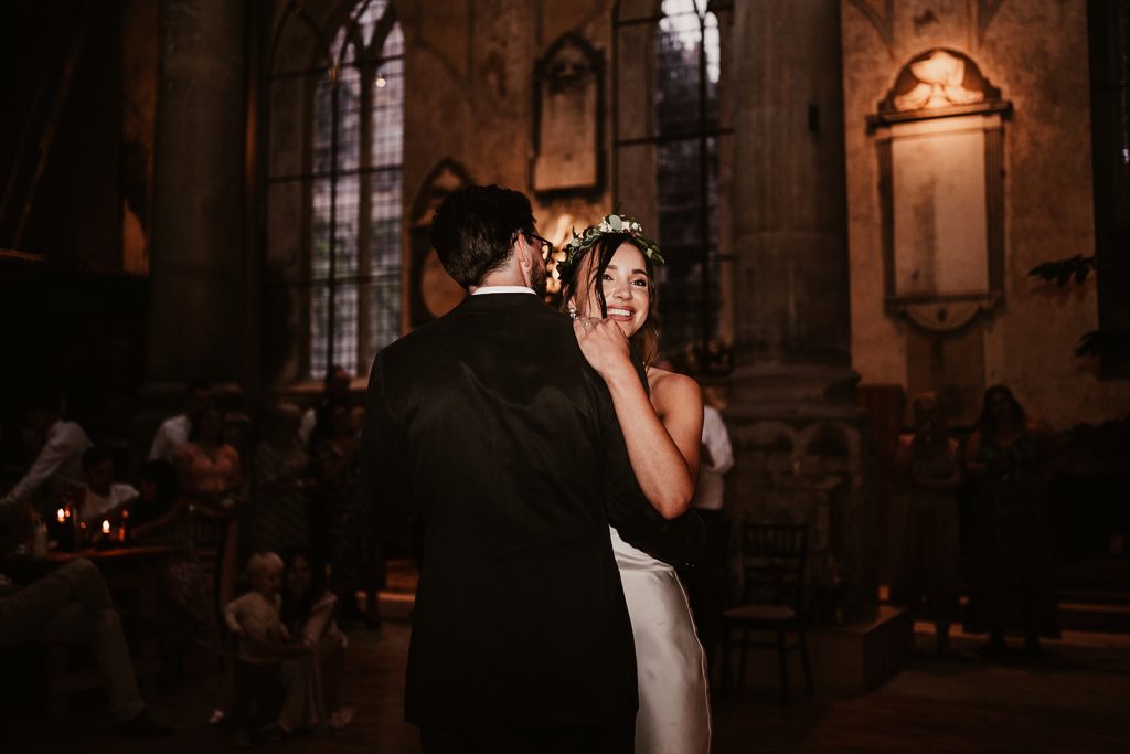 A newly married couple embrace on the dance floor of their Bristol wedding venue The Mount Without. The Groom's back is visible and the bride's face which is beaming with a huge smile. She wears her brown hair in a loose up-do accentuated with fresh flowers.