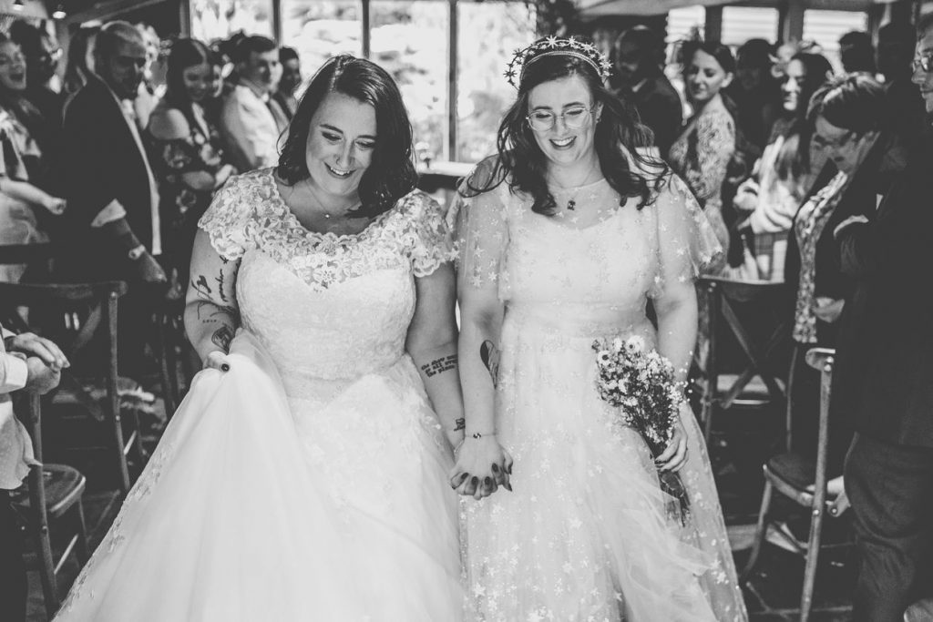 Two brides walk back down the aisle holding hands with beaming smiles on their faces as they become newly weds after their civil ceremony. The black and white picture shows both brides wearing wedding dresses, one in a princess style cut and the other with a celestial theme with stars covering the dress. This bride wears a delicate corwn on top of her head and holds a small bouquet of dried flowers.