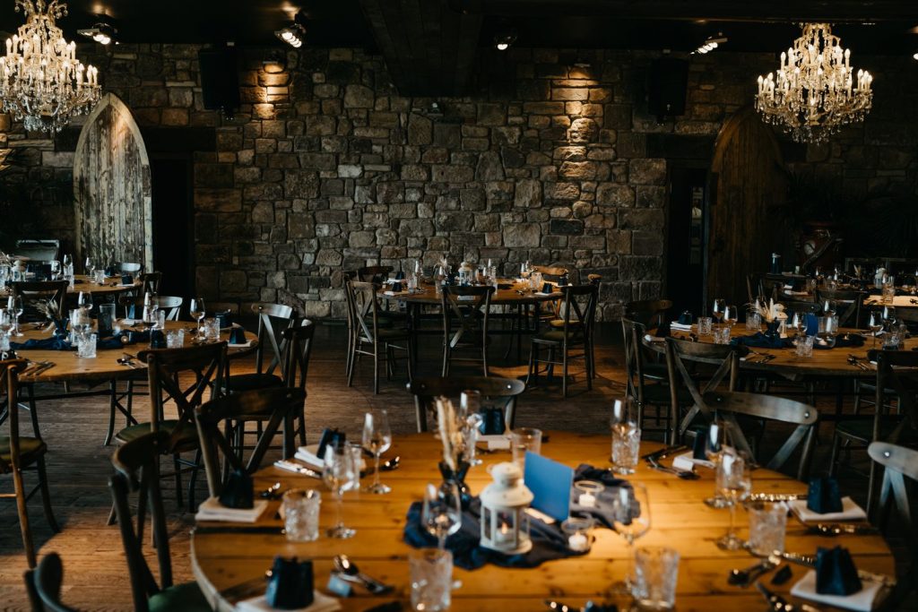 Round wooden tables with wooden chairs are decorated with blue and gold decor ready for a wedding breakfast. The styling is romantic and celestial yet quite minimal with barely any wedding flowers, creating a romantic, warm feel to the space.