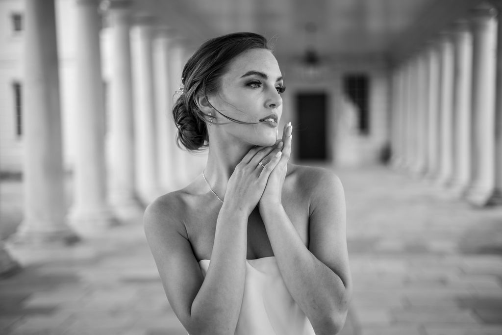 An editorial style image in black and white of a bride gazing longingly into the distance whilst she holds her hands together in a pose underneath her chin. The minimalism of her dress shows off her jewellery which features re-worked earrings from bridal designer Sarah Roberts London which are created from old vintage pieces.