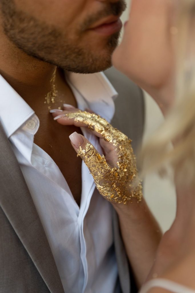 A couple share a tender kiss on the lips. The man wears an open collar white shirt with a grey jacket on top. The woman has a white vest top with thin spaghetti straps. One of her hands rests on the man's chest and neck and is painted with gold leaf.