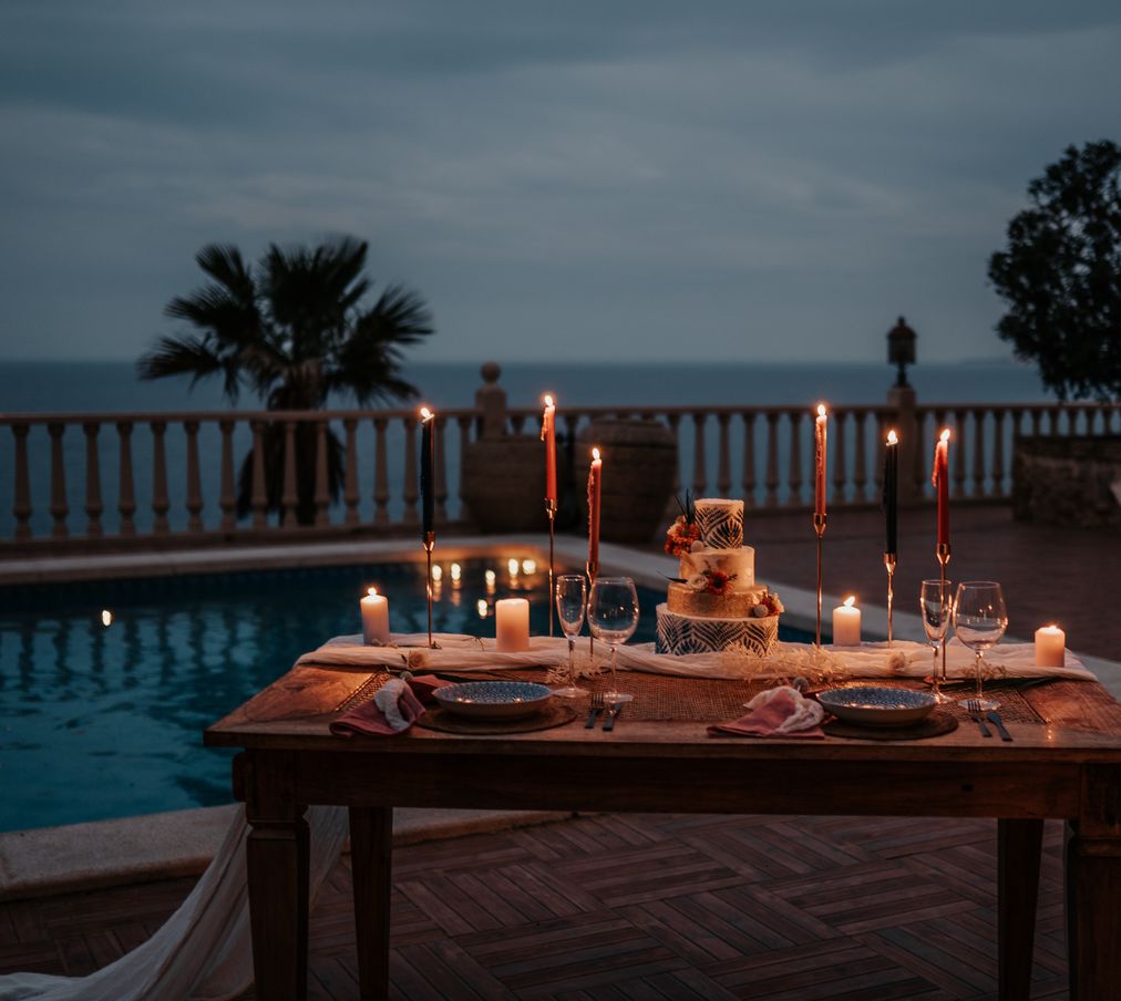 An intimate destination wedding tablescape is set out next to a private pool on the balcony of a Spanish villa. A textured fabric cloth runs the entire length of the table whilst tall tapered candles are placed delicately around the crockery and cutlery. The evening sky is jet blue due to the magical dusk hour and the table is romantic and warm with the light from the candles.