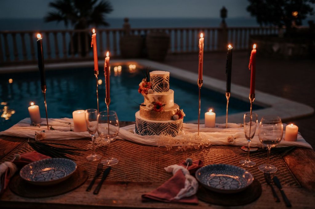 An intimate destination wedding tablescape is set out next to a private pool on the balcony of a Spanish villa. A textured fabric cloth runs the entire length of the table whilst tall tapered candles are placed delicately around the crockery and cutlery. The evening sky is jet blue due to the magical dusk hour and the table is romantic and warm with the light from the candles.
