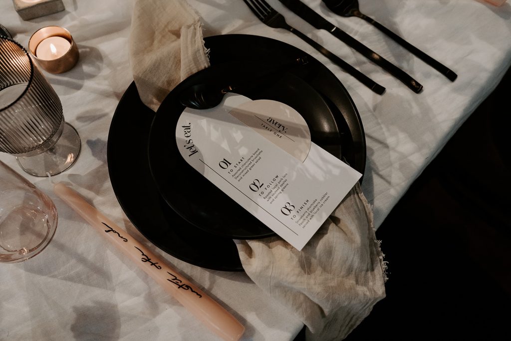 Modern wedding stationery on shades of cream sit on top of a hand dyed, textured piece of fabric which is set upon a black charger plate. Black cutlery is arranged to the right of the place setting and to the left is a cream tapered candle with hand writing in black along the length of the candle.