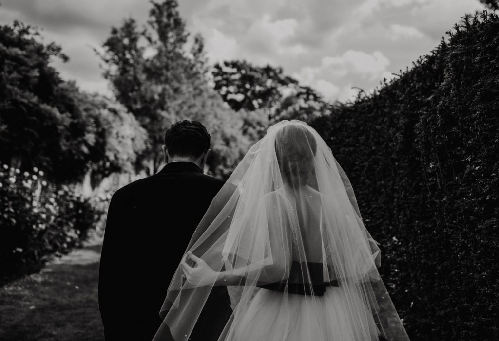 A black and white image of a bride and groom walking arm in arm as captured from above. The bride and groom hold each other around the waist. The groom wears a smart black jacket and the bride has a relaxed low bun underneath a long sheer veil.