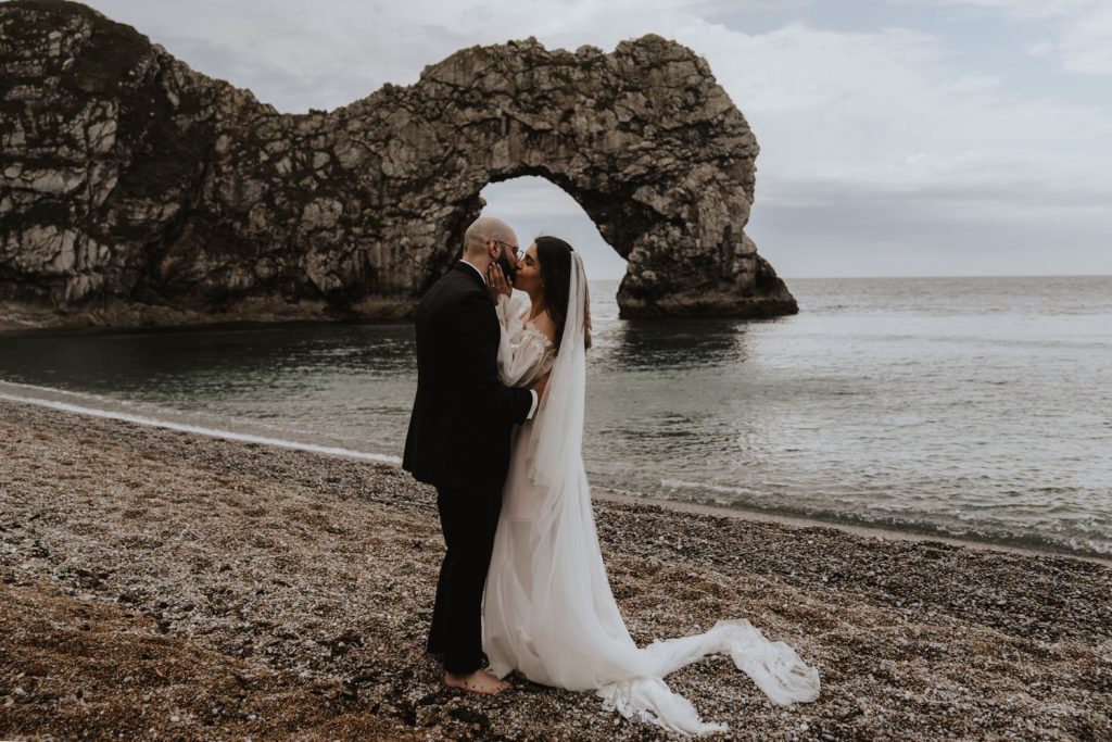 A bride and groom kiss one another whilst standing barefoot on a shingle beach at Durdle Door in Dorset. They hold one another with their hands as they embrace passionately. The bride wears a floaty bohemian style bespoke wedding dress with detachable billowing sleeves and a long Cathedral length veil.