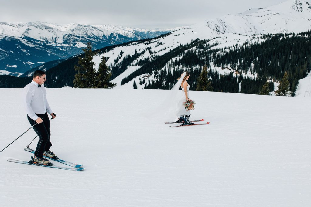 A bride wearing a sleeveless wedding dress and a long flowing veil skis next to her husband who wears a smart shirt and bow tie with trousers, down a snowy ski slope. In the background are large snow covered mountains
