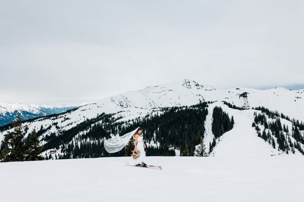 A bride wearing a sleeveless wedding dress and a long flowing veil skis down a snowy ski slope. In the background are large snow covered mountains
