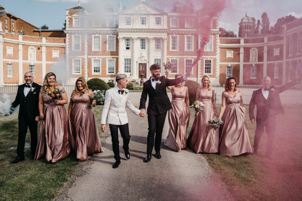 Two grooms stand hand in hand against the stately grand backdrop of Hawkstone Hall, their countryside wedding venue. One of the grooms and some of their wedding party hold smoke bombs which have pink and white smoke billowing out from them. The wedding party wear matching pink bridesmaid dresses.