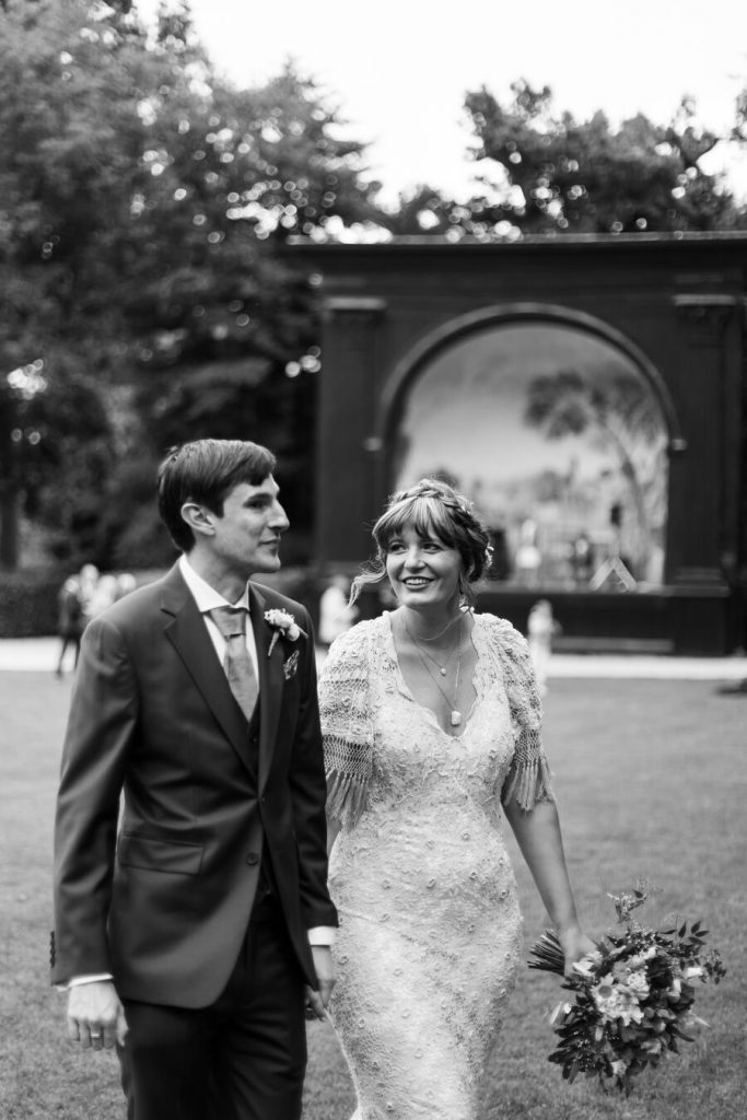 Black and white image of a bride and groom strolling around the estate at Larmer Tree, their wedding venue. The bride wears a vintage lace dress with short fringed sleeves and looks longingly at her husband with such adoration. The groom is looking away from his bride, with a happy smile on his lips.