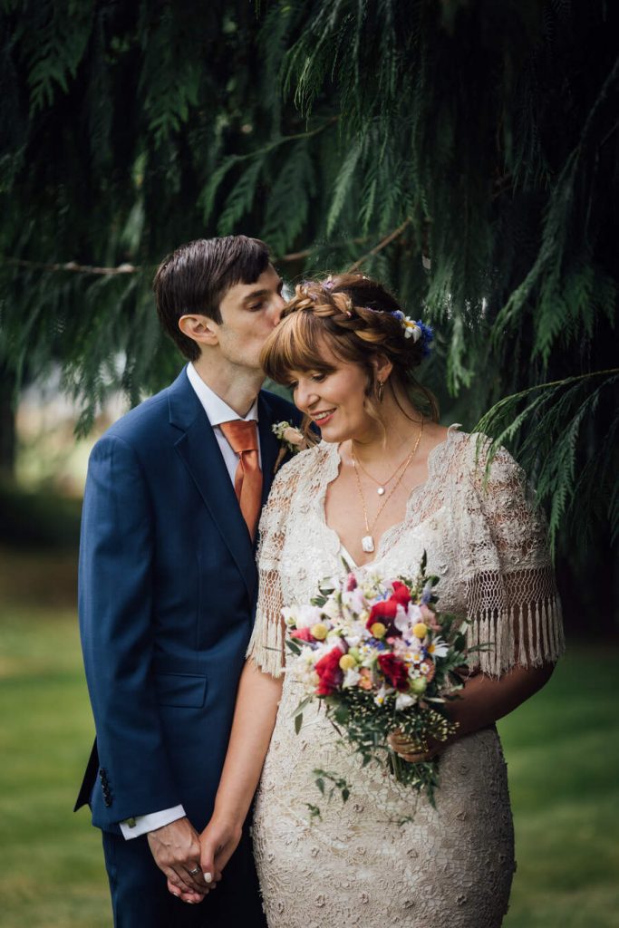 An intimate moment for a newly married bride and groom on their wedding day. The groom closes his eyes and gently kisses his new bride on the forehead; she smiles. The bride wears her hair up with a fringe and loose soft curls frame her face. Her dress is unique, made of vintage lace with short fringed sleeves.