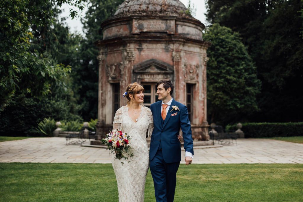 A newlywed couple post for portraits in front of the historic Roman Temple at Larmer Tree. The bride wears a vintage lace wedding dress with fringed sleeves and holds a colourful wedding bouquet.