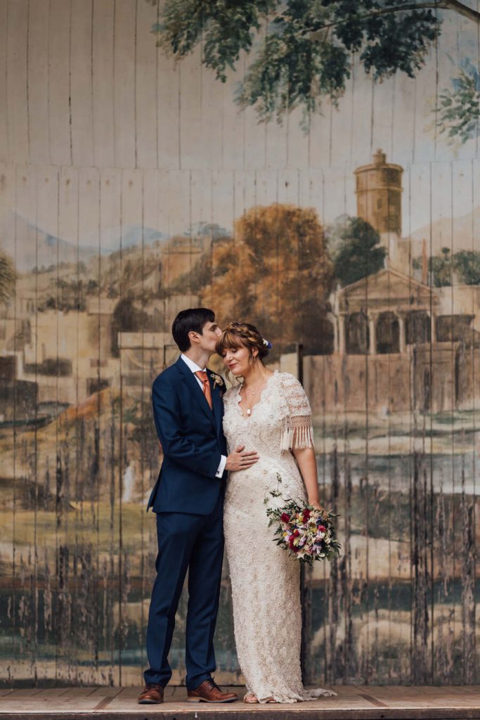 An intimate moment for a newly married bride and groom on their wedding day. The groom closes his eyes and gently kisses his new bride on the forehead; she smiles. The bride wears her hair up with a fringe and loose soft curls frame her face. Her dress is unique, made of vintage lace with short fringed sleeves.