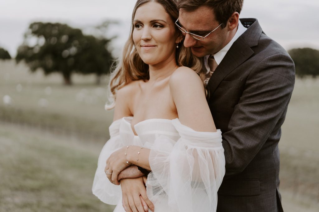 A newly married couple spend some quiet alone time at their countryside venue Wilderness Reserve to soak in their just married status. The groom stabnds behind his wife with his arms gently around her wait as he nuzzles into her neck. The bride smiles as she holds her hands over his.