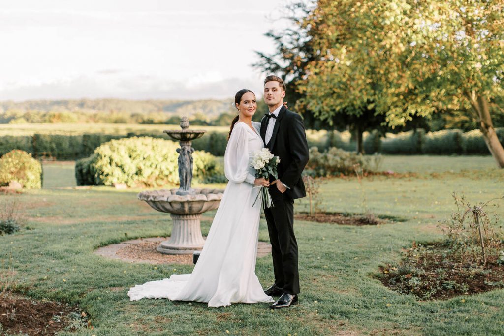 A couple stand facing the camera during their wedding portrait session. The groom looks sharp in a smart dark suit and bow tie whilst the bride has a modern feel, wearing a billowing long white wedding dress with full bishop style sleeves. She holds a small white bouquet in front of her and wears her brunette hair sleeked back into a low ponytail.