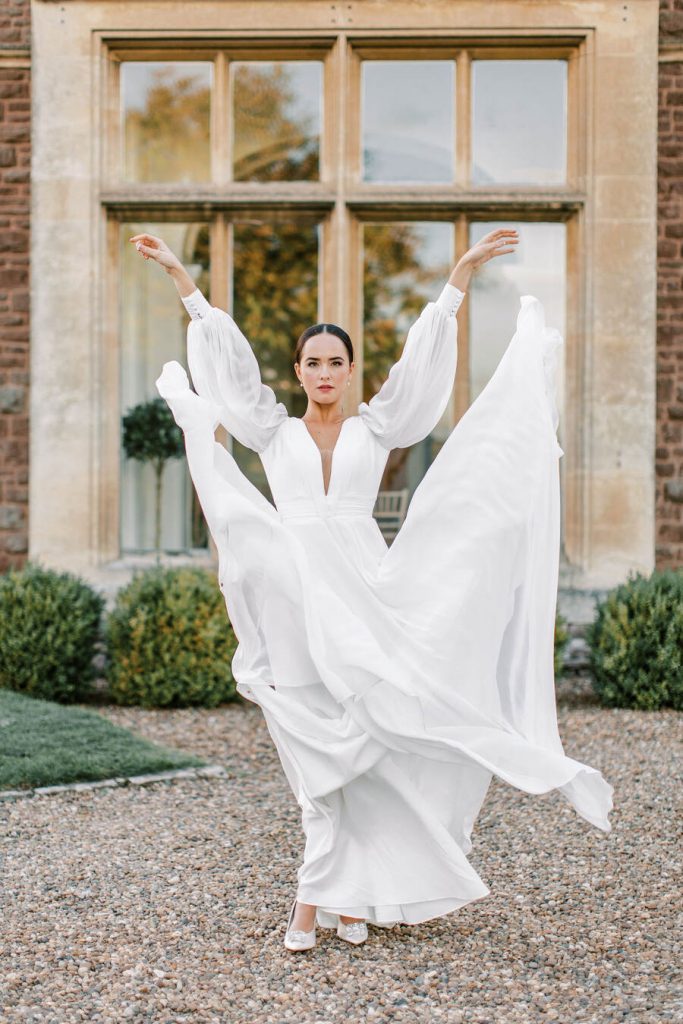 A bride stands outside her wedding venue wearing a full sleeve long airy white wedding dress. She lifts the skirt part of her dress up in the air and it falls in a billowing motion whilst her hands remain in the air.