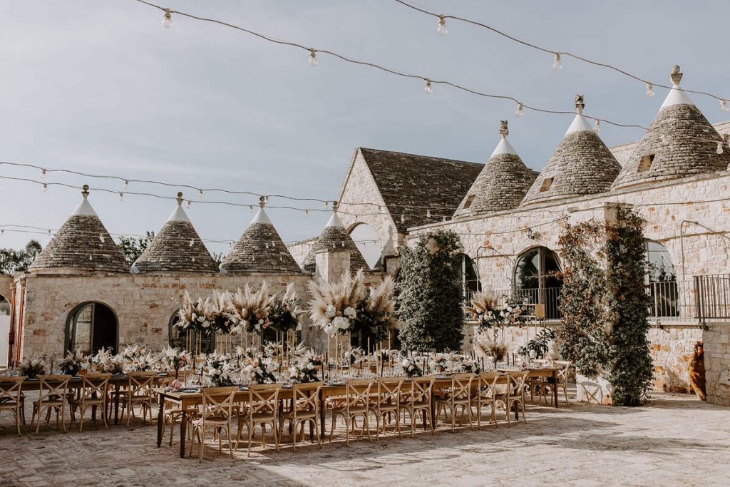 A beautiful Italian courtyard is set for a wedding with three long tables and on long top table. The day sky is a light blue and the tables are decorated in shades of coppers, blush and soft greys with lots of tall pampas floral displays lining the tables.