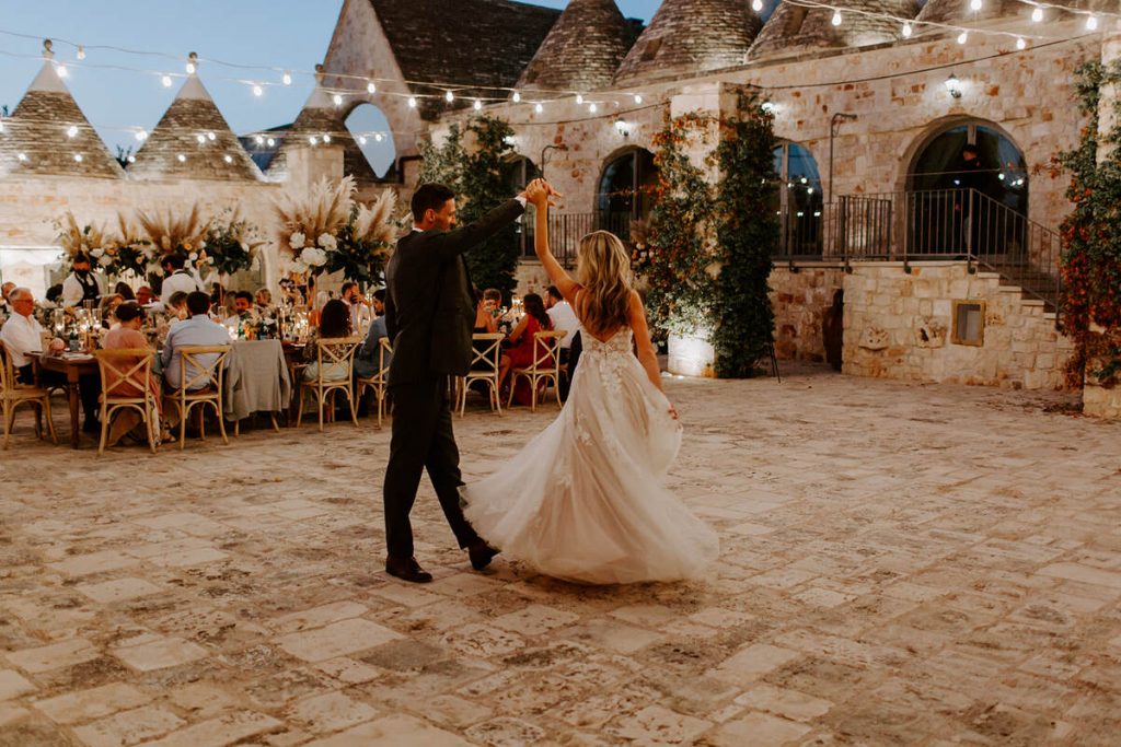 A newlywed couple take to the dance floor as the groom spins his bride around as they hold hands in the air. The brides large train on her wedding dress swishes beautifully as she twirls. The dance under the blue hour skies, in their outdoor courtyard wedding setting in Puglia. Behind them, long wedding tables can be seen, where their guests sit and chat as the night draws in.