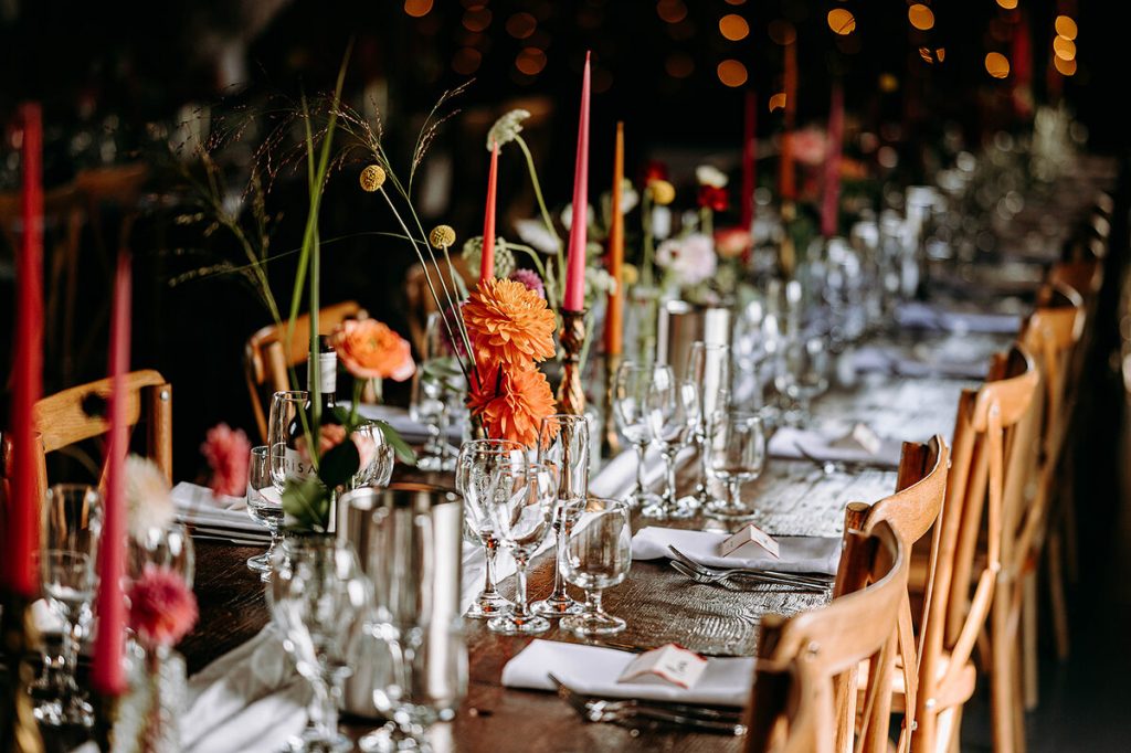 A wedding barn set up with décor and styling with a colourful, festival vibe. Long wooden tables are laden with flowers, crockery, cutlery and glassware with wooden chairs and colourful streamer banners hanging overhead.